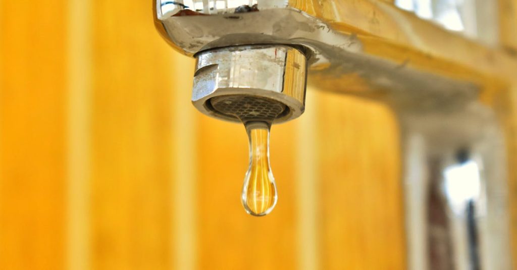 A close-up shot of a water droplet hanging from a stainless steel faucet, highlighting surface tension.